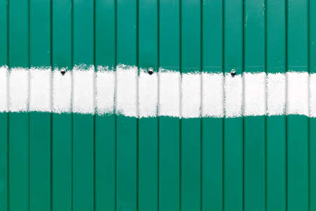 Green metal fence with vertical corrugated stripes, with a horizontal stripe of white paint in the middle and with three boltsの写真素材