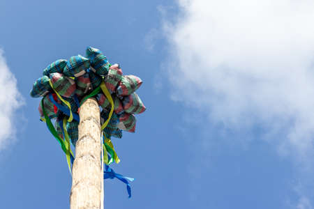 A wooden post, at the top of which are attached full bags of gifts and colorful ribbons against a blue sky. Traditional holiday entertainment by climbing a pole to get a giftの写真素材