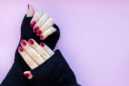 Female hands in black knitted gloves mitts with beautiful glossy manicure - burgundy, dark red color nails on a pink background with copy spaceの写真素材