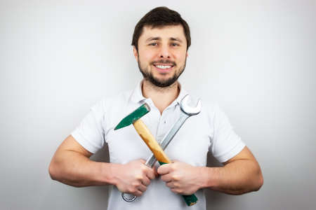 A smiling happy bearded man in a white t-shirt with a hammer and a wrench, holding them crosswise