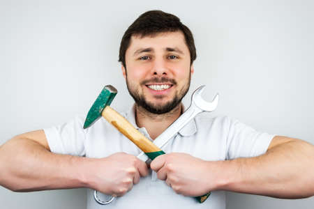 A smiling happy bearded man in a white t-shirt with a hammer and a wrench, holding them crosswiseの写真素材
