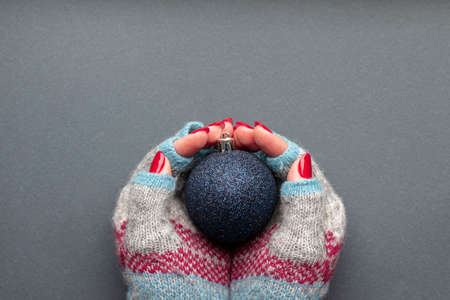 Female hands in warm winter knitted gloves with patterns, with dark blue shiny Christmas ball and with beautiful manicure - red glittered nails on gray background with copy spaceの写真素材