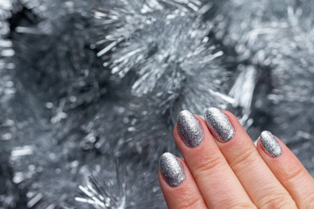 Female hand with beautiful holiday manicure - silver glittered nails on Christmas tinsel background. Selective focus. Closeup view. Blurred background with copy spaceの写真素材