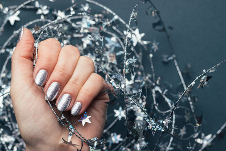 Female hand with beautiful holiday manicure - silver glittered nails with Christmas twisted wire with stars. Selective focus. Closeup view. Blurred gray background with copy spaceの写真素材