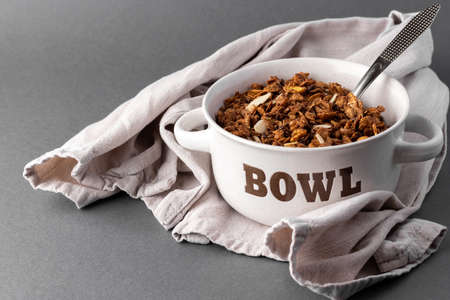 Organic homemade roasted granola cereal with oats, flax, almonds, chocolate in white round glass bowl with metal spoon and linen textile towel on gray background. Natural muesli breakfastの写真素材