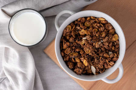 Organic homemade roasted granola cereal with oats, flax, almonds, chocolate in white round bowl with glass of milk on wooden tray or board on linen textile. Top view natural muesli breakfast, flat layの写真素材