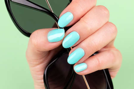 Female hand with beautiful manicure - mint blue nails with sunglasses on green background, closeup, selective focusの写真素材