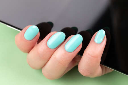 Female hand with beautiful manicure - mint blue nails with smartphone on green background. Selective...