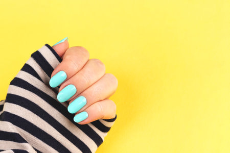 Female hand in sailor striped shirt with beautiful manicure - mint blue nails on yellow background with copy spaceの写真素材