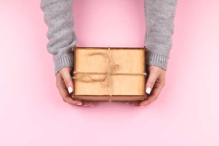Woman hands in gray knitted sweater with white nails holding wooden rustic gift box decorated with hemp rope on pink background. Top view, flat lay compositionの写真素材
