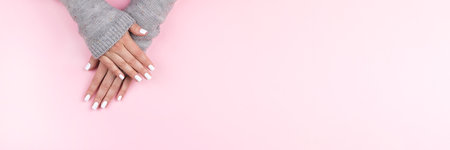 Female hands in gray knitted sweater with beautiful manicure - white nails on pink background, wide banner with copy space. Nail care conceptの写真素材