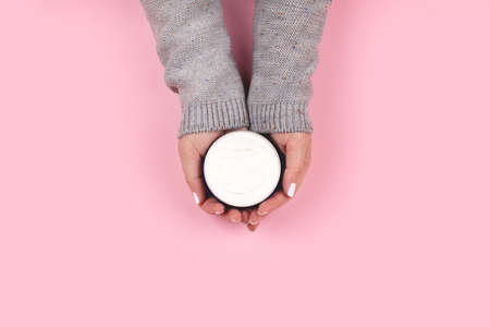 Female hands in gray knitted sweater with white nails manicure holding jar with cream on pink...