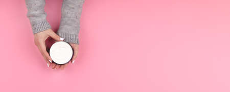Female hands in gray knitted sweater with white nails manicure holding jar with cream on pink...