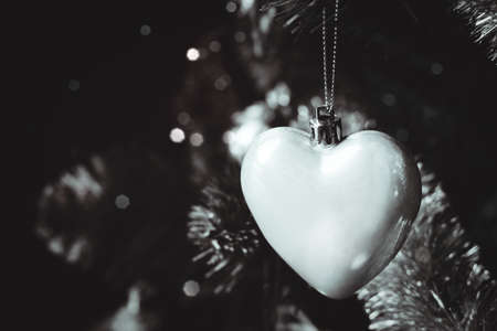 Silver gray glossy heart on Christmas tree with blurred lights bokeh. Selective focus. Holiday card with decorations for the New Year's Eve. Atmospheric black and white photoの写真素材