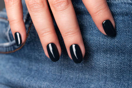Female hand with beautiful manicure - dark black nails on jeans denim textile, closeup fingers. Selective focusの写真素材