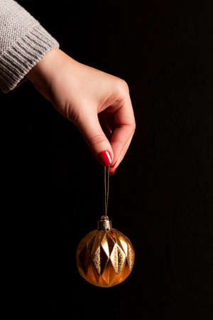 Female hand in gray knitted sweater with golden Christmas ball with beautiful manicure - red nails against black background. Nail and skin care conceptの写真素材