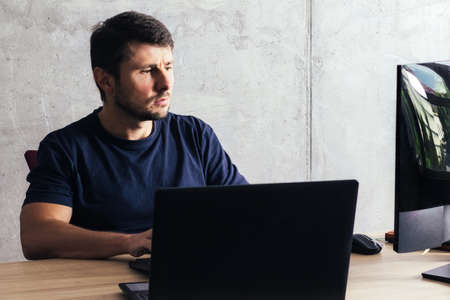 Handsome serious bearded man sitting at the office desk, working on laptop, looking at computer screen against background of concrete wall in modern officeの写真素材