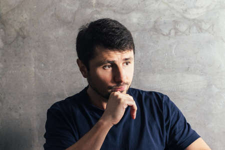 Handsome pensive serious bearded man with hand at chin sitting at the modern office against concrete...