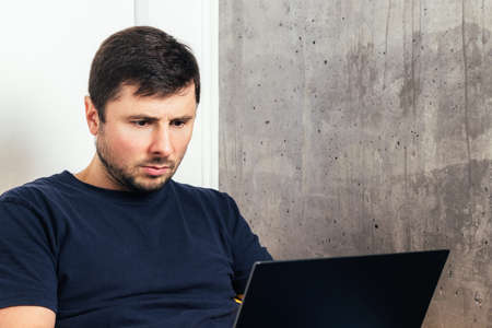 Handsome bearded man looking at his laptop screen against background of concrete wall in modern...