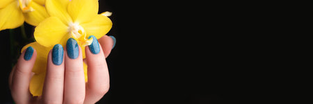 Female hand with beautiful manicure - blue glitter nails with yellow phalaenopsis orchid flower on black background with copy space banner. Wide panoramic headerの写真素材