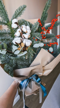 Handheld bouquet featuring cotton flowers, evergreen branches, and red berries, wrapped in brown paper with ribbon.の写真素材