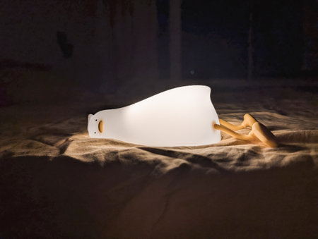 Modern minimalist lamp placed on bed with soft glow against dark background.の写真素材