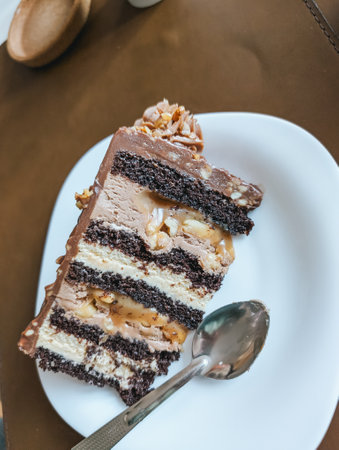 Layered chocolate cake slice on white plate with spoon. Rich dessert with creamy layers and nuts.