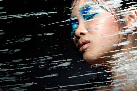 Beautiful Asian girl with bright blue makeup behind glass and drops of wax. Beauty face.の写真素材