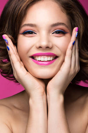 Beautiful model girl with bright makeup, smile and colored  nail polish. Beauty face. Short colorful nails. Picture taken in the studio on a pink background.の写真素材