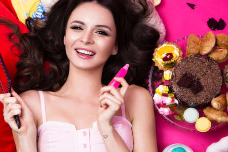 Beautiful fresh girl doll lying on bright backgrounds surrounded by sweets, cosmetics and gifts. Fashion beauty style. Photos shot in the studio.の写真素材