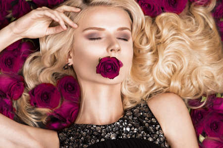Beautiful blond girl lying on a background of roses. Curls, red lipstick, evening dress. The beauty...