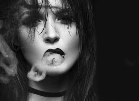 Beautiful sexy brunette girl with bright makeup, red lips, smoking with smoke from mouth. beauty face. Black and white. Photos shot in the studio on a black background.の写真素材
