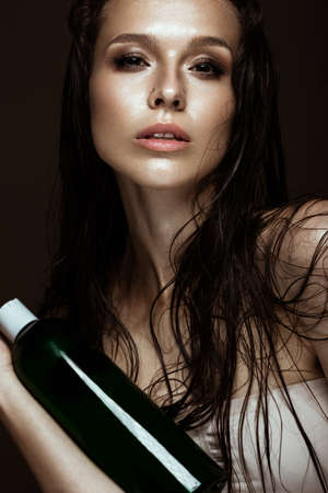 Beautiful girl with a bright make-up and wet hair and skin with bottle of cosmetic products in the hands. Beauty face. Picture taken in the studio on a black background.の写真素材