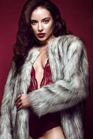 Beautiful sexy long-haired brunette woman in red lingerie and fur coat posing in the studio. The beauty of the face and body. Photos shot in a studioの写真素材
