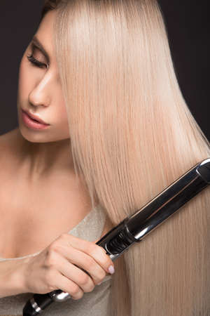 Beautiful blond girl with a perfectly smooth hair, classic make-up with curling styler in the hands. Beauty face. Picture taken in the studio.の写真素材