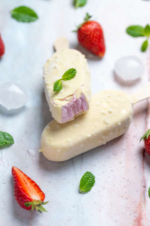 Strawberry ice cream on stick covered with white chocolate on marble table, flat lay