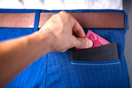 The person pulls a red packet of condom out of the wallet from his trouser pocket. Concept of contraception and prevention of sexually transmitted diseases, stop HIV.の写真素材