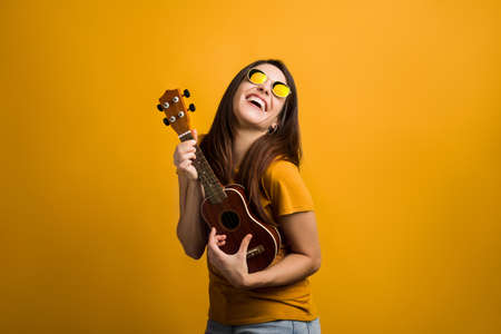 Attractive girl with long hair having fun with ukulele and modern glasses. Caucasian smiling girl enjoying music and posing on orange background.の写真素材