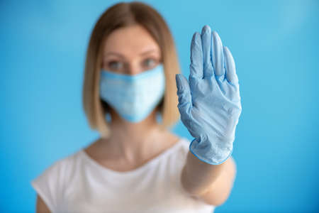 Attractive female nurse showing stop sign with his hand in glove gesture isolated on blue background. Medical concept of infectionの写真素材