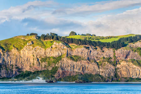 Rocky cliff face with bush and meadows on top at New Zealand coast near Dunedin at Otago Region Southern island - cloudy skyの写真素材