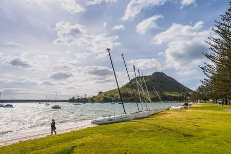 Tauranga, New Zealand - November 20, 2014: Mount Maunganui is extinct volcano that rises above the town of Tauranga. It is also known as the Mount. Adjacent to the Mount is a park where people relax. Sailing boat in the foreground in backlight.のeditorial素材