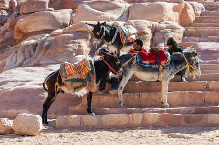Petra, Jordan - November 20, 2010: Young Arabian men sitting on stone steps and waiting for tourists in the ruins of ancient town Petra. Three donkey standing around them.のeditorial素材