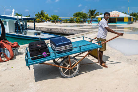 Gulhi Island, Maldives - November 17, 2017: Worker carries suitcases of tourists on a wheelbarrow, in the background boats, stone pier and Indian Ocean, tropical Gulhi Island, Maldives.のeditorial素材