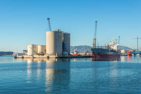 Malaga, Spain - December 4, 2018: Cement Carrier Vessel Glory Tellus and bulk storage of concrete cement silos and Bacardi tanks at entrance of Malaga Port, Andalusia, Spain.のeditorial素材