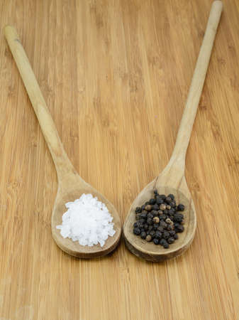 Sea salt crystals and black pepper in wooden spoons against a bamboo background の写真素材