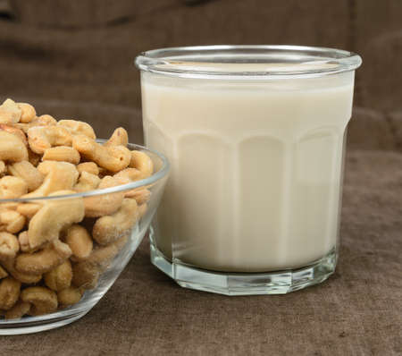 A glass of cashew milk with a bowl of cashews on a brown tablecloth.の写真素材