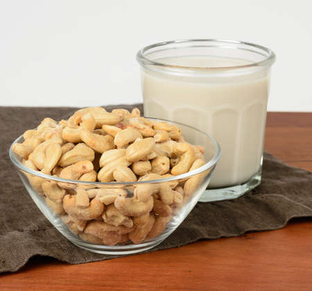 A glass of cashew milk with a bowl of cashews on a brown tablecloth.の写真素材