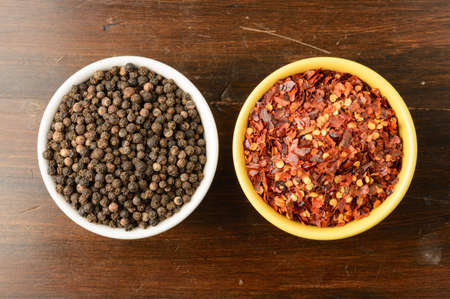 Black peppercorn in a white bowl next to crushed red pepper in a yellow bowl.の写真素材