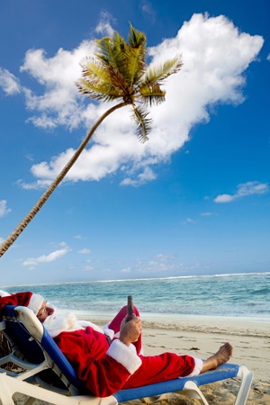 Santa claus is on vacation. He is resting on a sun lounger on exotic beach drinking a beer.の写真素材