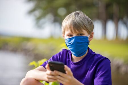 Young boy looks at camera, while texting, wearing a face mask for protection during coronavirus pandemicの写真素材
